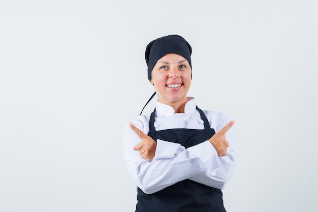 Cozinheira de uniforme, avental apontando para longe e olhando alegre, vista frontal.