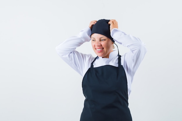 Cozinheira de uniforme, avental de mãos dadas na cabeça e olhando animada, vista frontal.