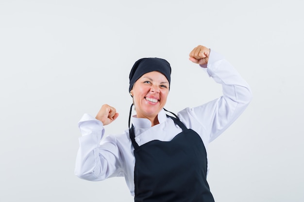 Foto grátis cozinheira de uniforme, avental, mostrando o gesto do vencedor e olhando feliz, vista frontal.