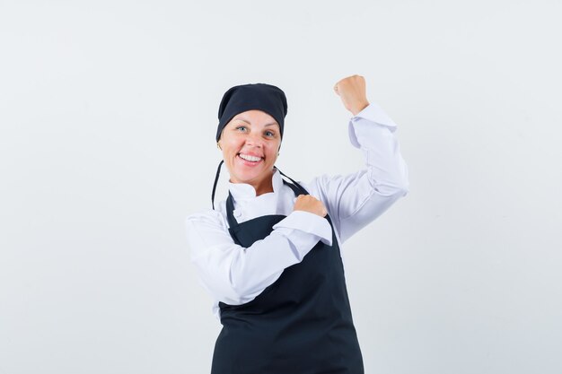 Cozinheira de uniforme, avental, mostrando o gesto do vencedor e parecendo feliz, vista frontal.