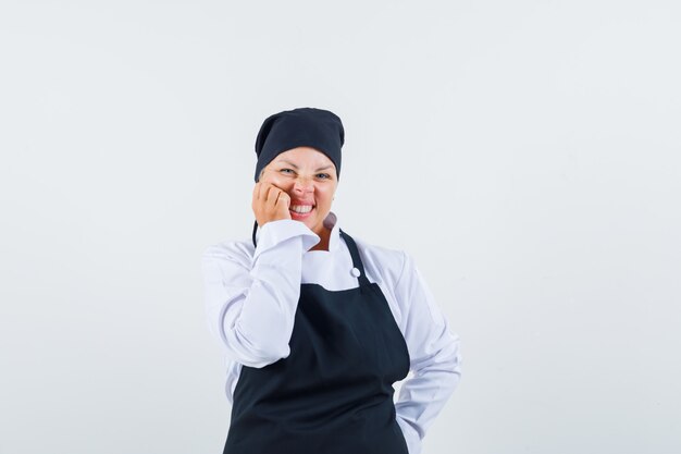 Cozinheira de uniforme, avental segurando a mão na bochecha e olhando bonito, vista frontal.
