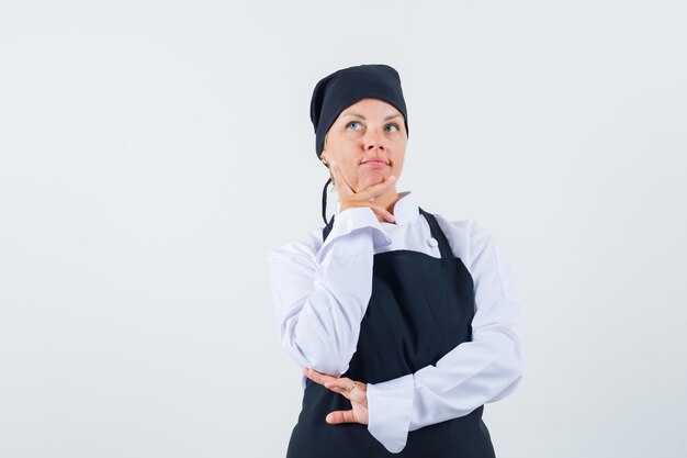 Cozinheira de uniforme, avental segurando a mão no queixo e olhando pensativa, vista frontal.