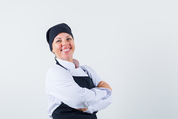 Cozinheira em pé com os braços cruzados no uniforme, avental e parecendo feliz, vista frontal.