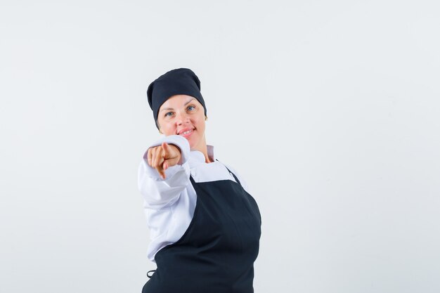 Cozinheira feminina, apontando para a câmera de uniforme, avental e parecendo confiante, vista frontal.