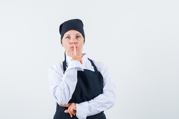 Cozinheira feminina mostrando gesto de silêncio no uniforme, avental e olhando cuidadosa, vista frontal.