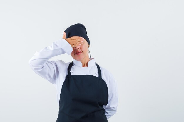 Cozinheira feminina segurando a mão nos olhos de uniforme, avental e parecendo calma, vista frontal.