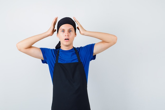 Cozinheiro adolescente masculino com as mãos perto da cabeça, abrindo a boca na camiseta, avental e olhando horrorizado, vista frontal.