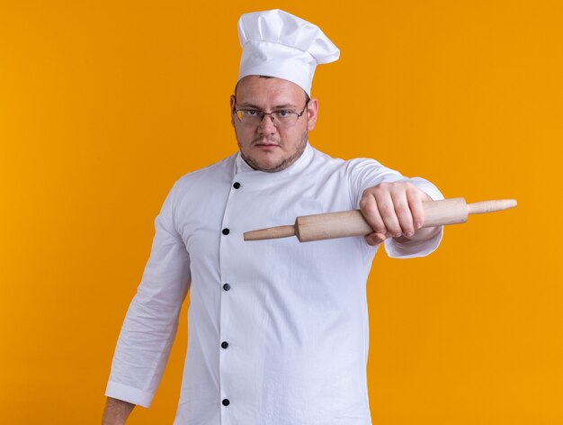 Foto grátis cozinheiro adulto confiante usando uniforme de chef e óculos, olhando para a câmera, esticando o rolo de massa em direção à câmera, isolada em um fundo laranja