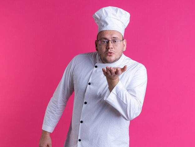 cozinheiro adulto confiante usando uniforme de chef e óculos olhando para a frente enviando beijo de golpe isolado na parede rosa