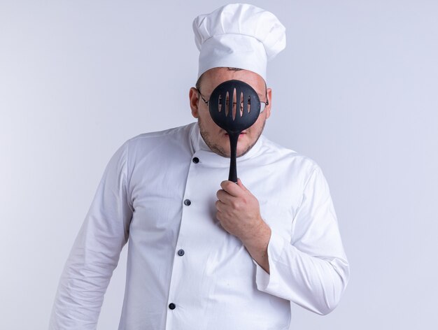 Cozinheiro adulto impressionado usando uniforme de chef e óculos segurando uma escumadeira na frente do rosto através dela isolada na parede branca