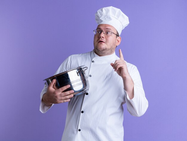 cozinheiro adulto impressionado usando uniforme de chef e óculos segurando uma panela, olhando para o lado apontando para cima, isolado na parede roxa