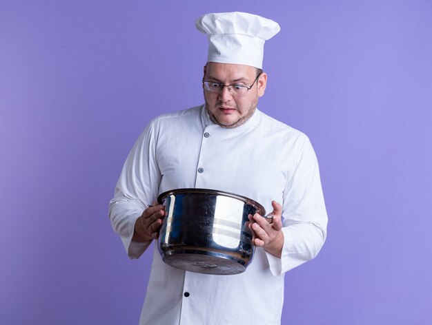 cozinheiro adulto surpreso usando uniforme de chef e óculos segurando uma panela, olhando para dentro, isolada na parede roxa com espaço de cópia