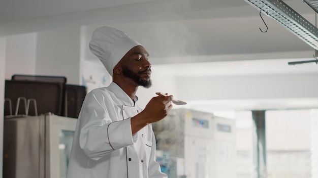 Cozinheiro americano africano provando sopa de legumes no fogão, sentindo-se confiante sobre o prato de cozinha profissional. chef autêntico cozinhando refeições frescas e comida deliciosa na cozinha do restaurante.