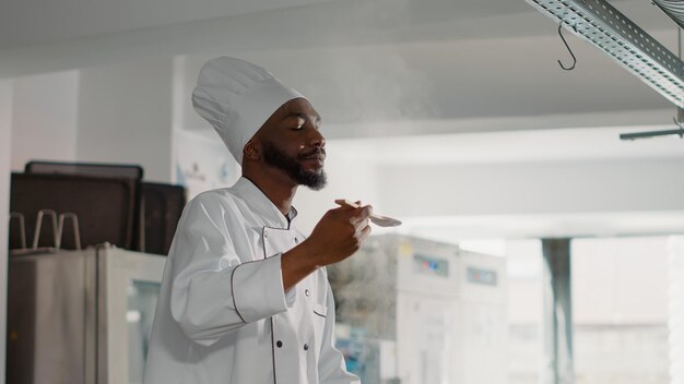 Cozinheiro americano africano provando sopa de legumes no fogão, sentindo-se confiante sobre o prato de cozinha profissional. Chef autêntico cozinhando refeições frescas e comida deliciosa na cozinha do restaurante.