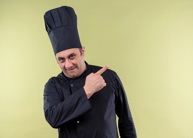 Cozinheiro chef masculino vestindo uniforme preto e chapéu de cozinheiro olhando para a câmera sorrindo alegremente apontando com o dedo indicador para o lado em pé sobre fundo verde