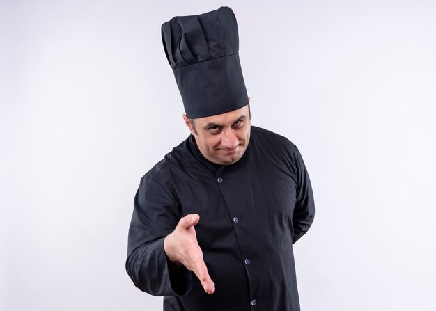 Cozinheiro chef masculino vestindo uniforme preto e chapéu de cozinheiro, olhando para a câmera, sorrindo amigável, oferecendo hnad em pé sobre um fundo branco