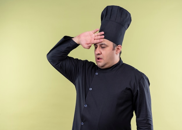 Cozinheiro chef masculino vestindo uniforme preto e chapéu de cozinheiro parecendo cansado e sobrecarregado com a mão na cabeça em pé sobre um fundo verde