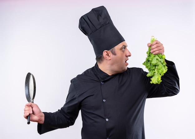 Cozinheiro chef masculino vestindo uniforme preto e chapéu de cozinheiro segurando alface fresca e uma panela parecendo de lado surpreso em pé sobre um fundo branco