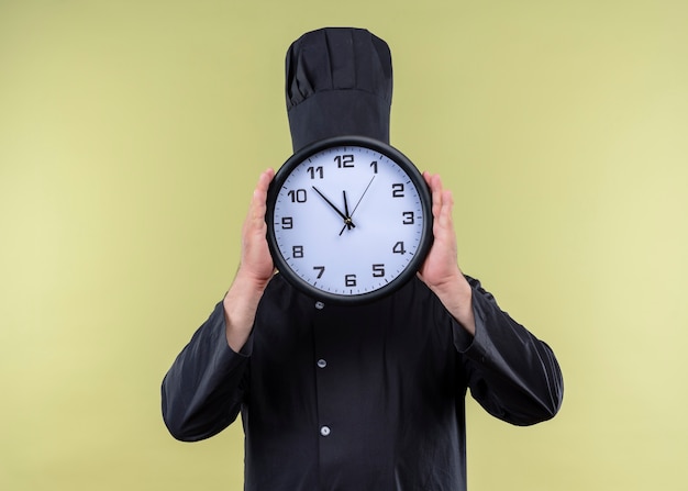 Cozinheiro chef masculino vestindo uniforme preto e chapéu de cozinheiro segurando um relógio de parede, escondendo o rosto atrás dele, em pé sobre um fundo verde