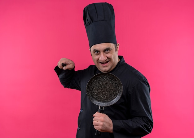 Cozinheiro chef masculino vestindo uniforme preto e chapéu de cozinheiro segurando uma panela, ameaçando com uma faca, olhando para a câmera com um sorriso no rosto em pé sobre um fundo rosa