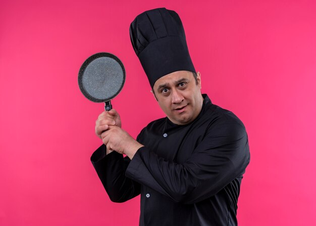 Cozinheiro chef masculino vestindo uniforme preto e chapéu de cozinheiro segurando uma panela, olhando para a câmera confuso em pé sobre um fundo rosa