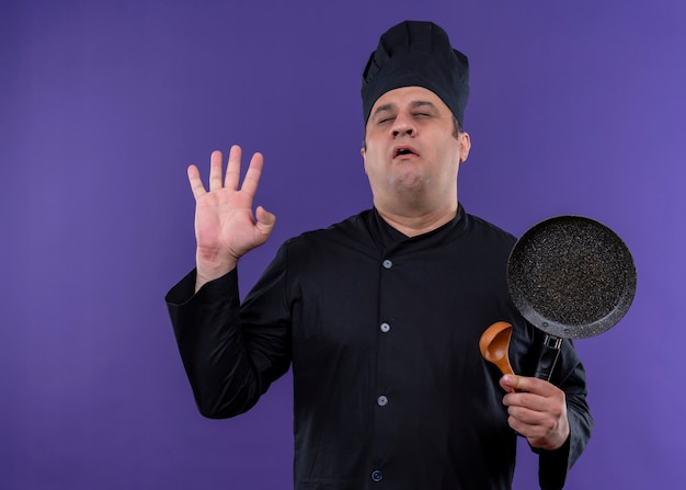 Foto grátis cozinheiro chefe masculino preocupado, vestindo uniforme preto e chapéu de cozinheiro, segurando uma frigideira com os olhos fechados, de pé sobre um fundo roxo