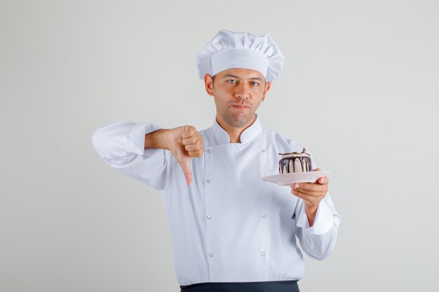 Cozinheiro chefe masculino que guarda o bolo e que mostra o polegar para baixo na vista dianteira do uniforme, do avental e do chapéu.