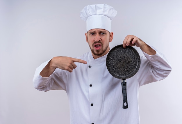 Cozinheiro chefe profissional masculino decepcionado com uniforme branco e chapéu de cozinheiro segurando uma panela apontando com o dedo para ela, de pé sobre um fundo branco