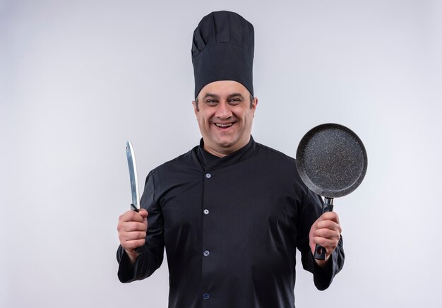 Cozinheiro de meia-idade sorridente com uniforme de chef segurando uma frigideira e um cutelo com espaço de cópia