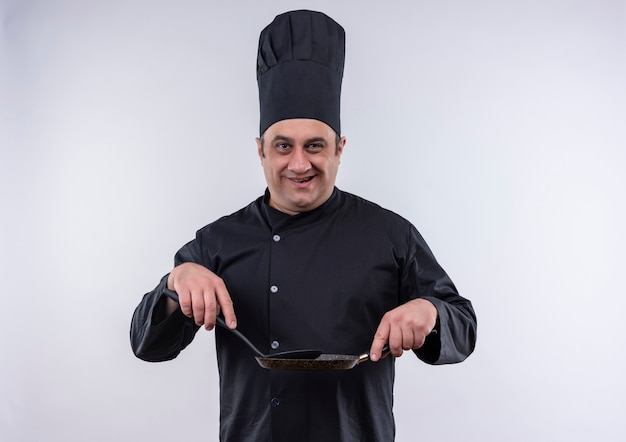 Cozinheiro de meia-idade sorridente com uniforme de chef segurando uma frigideira e uma espátula