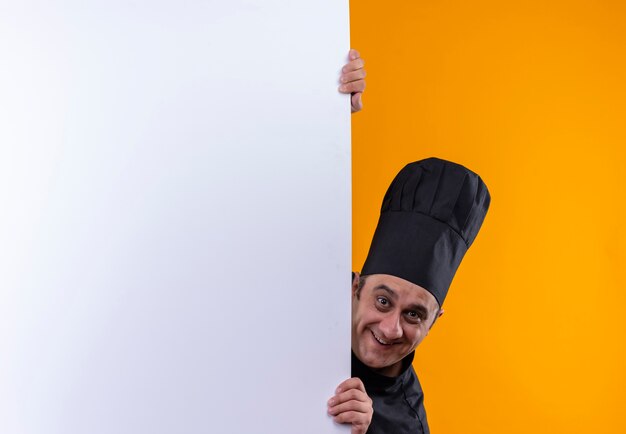 Cozinheiro de meia-idade sorridente com uniforme de chef segurando uma parede branca sobre fundo amarelo com espaço de cópia