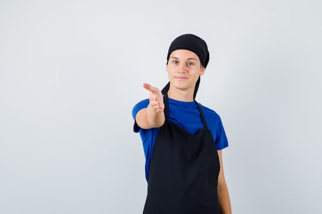 Cozinheiro jovem adolescente oferecendo aperto de mão como saudação em t-shirt, avental e olhando alegre. vista frontal.