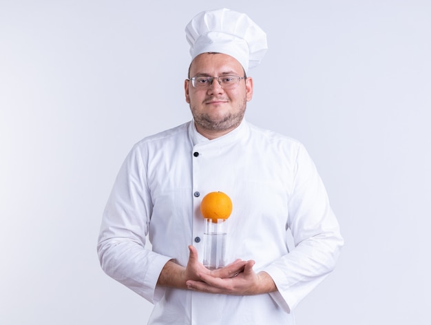 Foto grátis cozinheiro masculino adulto satisfeito vestindo uniforme de chef e óculos segurando um copo de água com laranja, olhando para frente, isolado na parede branca