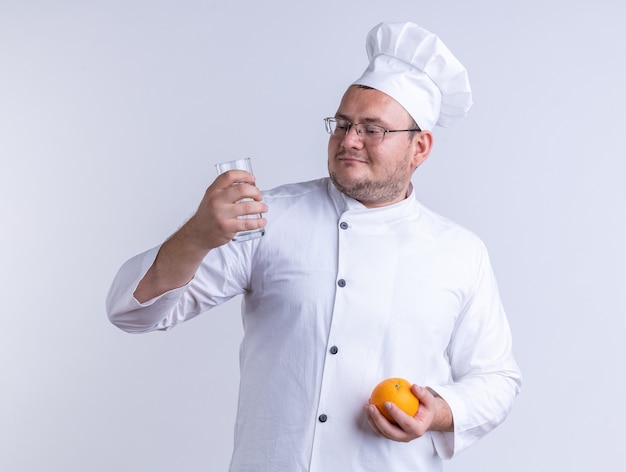 Cozinheiro masculino adulto satisfeito vestindo uniforme de chef e óculos segurando uma laranja e um copo de água olhando para um copo de água isolado na parede branca