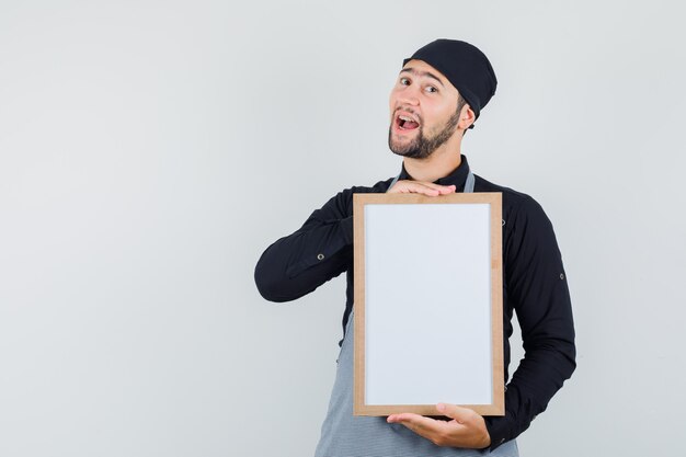 Cozinheiro masculino segurando quadro branco na camisa, avental e olhando alegre, vista frontal.