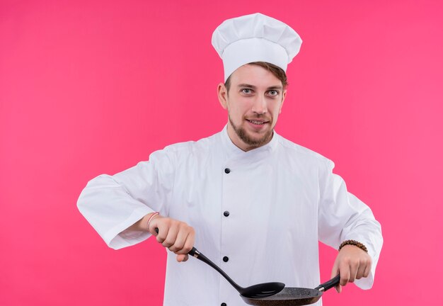 Cozinheiro olhando para o sorriso da câmera no rosto com a colher na panela em pé sobre a parede rosa