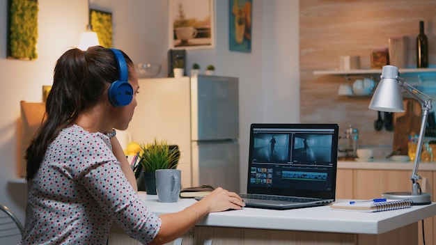 Criador de conteúdo freelancer trabalhando horas extras de casa para respeitar o prazo. Mulher cinegrafista editando montagem de filme de áudio em laptop profissional sentada na mesa de uma cozinha moderna à meia-noite