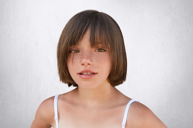 Criança adorável com olhos castanhos encantadores, pele sardenta e lábios finos, com um penteado elegante, vestindo roupas de verão, olhando diretamente para a câmera enquanto posava em branco.