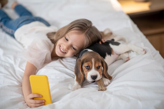 Criança alegre do sexo feminino fotografando a si mesma e ao cachorrinho com um brinquedo nos dentes