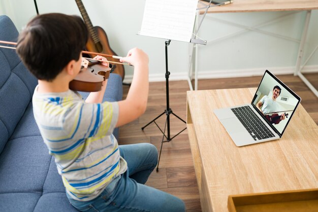 Criança com habilidades musicais tocando violino em casa enquanto presta atenção ao seu professor de música em uma videochamada