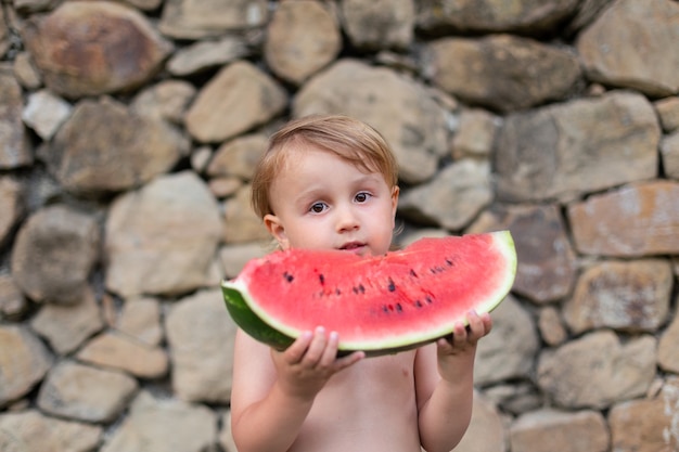 Criança comendo melancia no jardim durante as férias de verão. as crianças comem frutas ao ar livre. lanche saudável para crianças. pequeno bpy brincando no jardim segurando uma fatia de melancia.