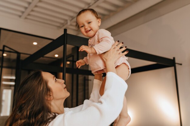 Criança de blusa bege fica feliz que sua mãe a joga para cima, brincando com ela em um apartamento aconchegante.