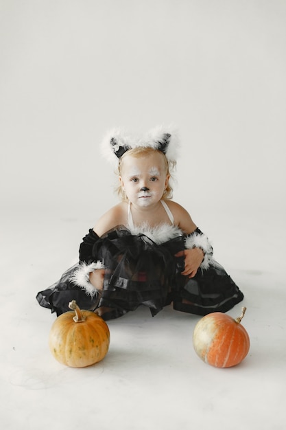 Criança de menina vestida de vestido preto como um gato sentado perto de duas abóboras. menina com o rosto pintado como o rosto de um gato