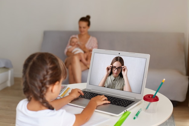 Criança do sexo feminino de cabelos escuros com rabo de cavalo vestindo camiseta branca, sentada na frente da tela do laptop, tendo aula online, olhando e ouvindo seu professor.