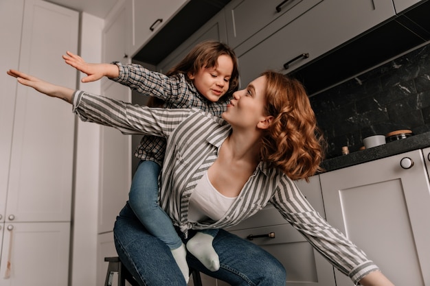 Criança do sexo feminino se senta nas costas das mães e brinca com ela como um avião na cozinha.