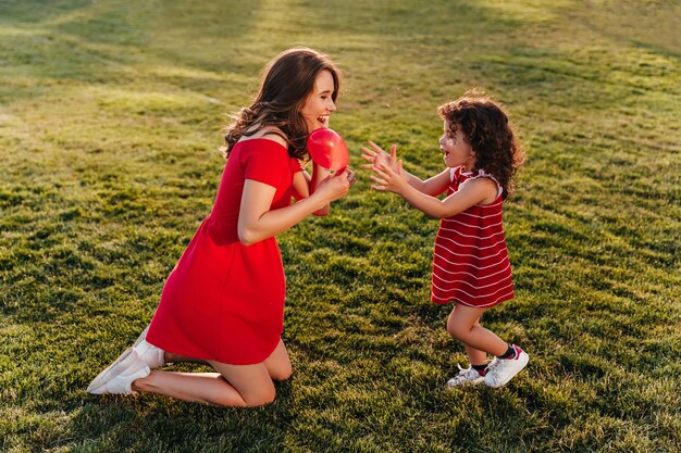 Criança engraçada no vestido vermelho enjoyng dia de verão com a mãe. Foto ao ar livre de uma linda mulher morena brincando com a filha na grama.