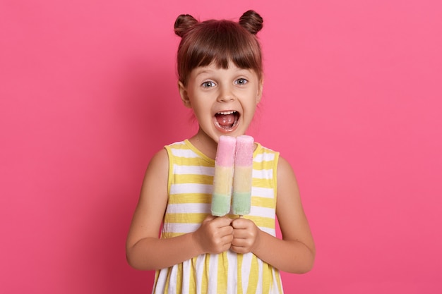 Criança europeia pequena mordendo sorvete de frutas, menina encantadora com a boca bem aberta, vestindo roupas de verão, parece feliz, se divertindo com uma sobremesa deliciosa.