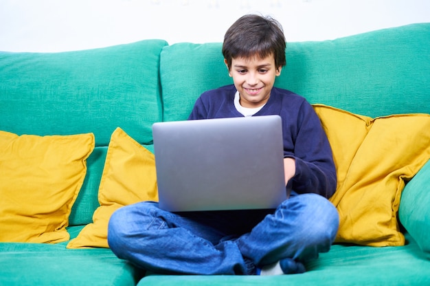 Criança inteligente e alegre usando laptop sentado no sofá em casa.