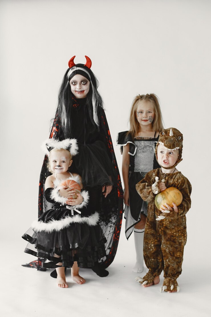 Foto grátis criança menino vestido com fantasia marrom de um dinossauro segurando uma abóbora. meninas vestidas de preto se vestem como um gato e como uma bruxa