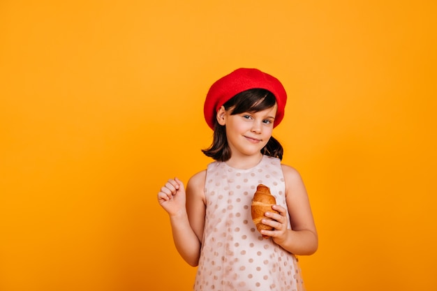 Criança morena curiosa posando na parede amarela. menina pré-adolescente comendo croissant.